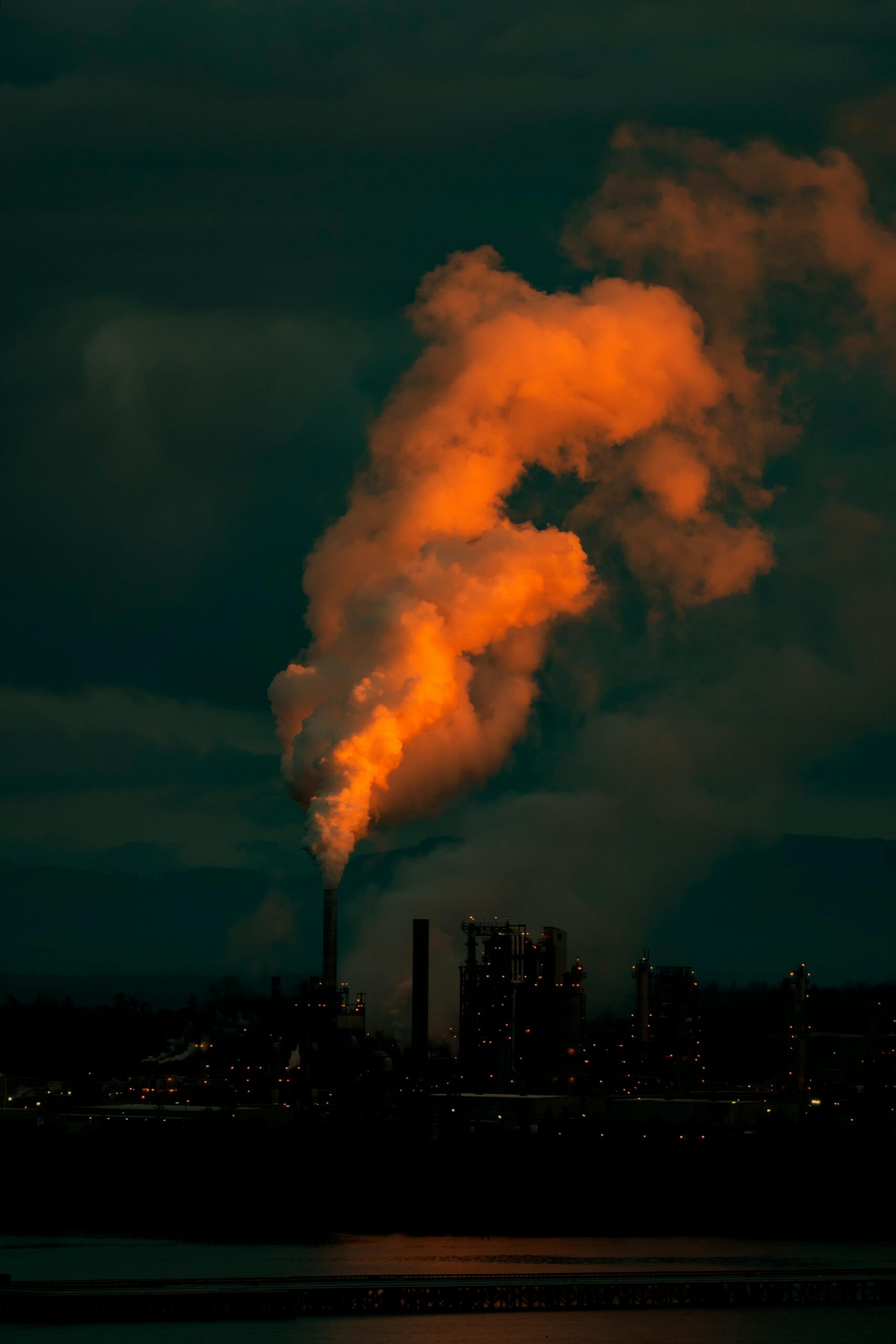 Bright smoke from smokestack top creating thick wavy cloud in twilight in town under dark sky illuminated by artificial light